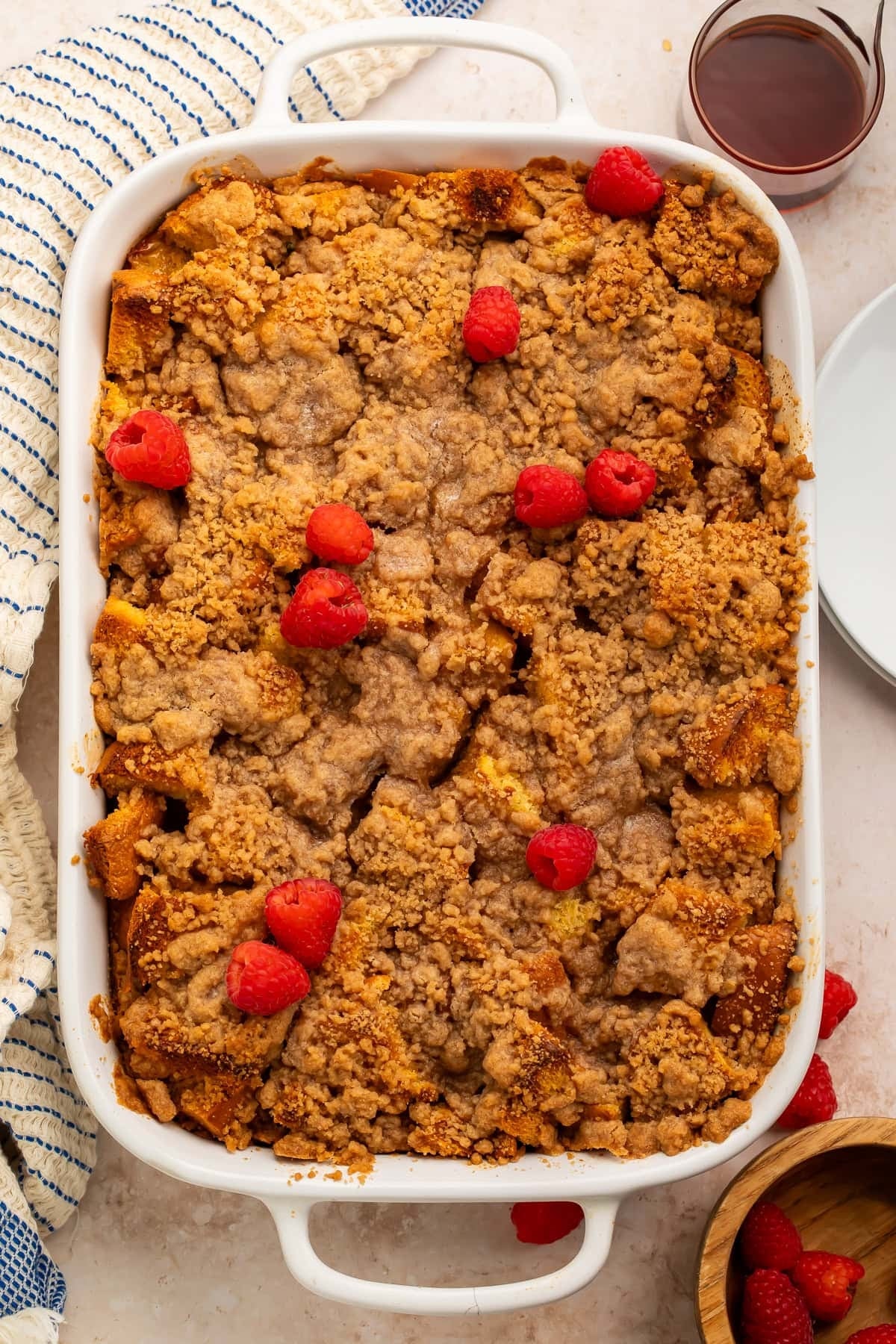 Baked crumble dessert topped with fresh raspberries in a white dish, next to a drink and empty plates