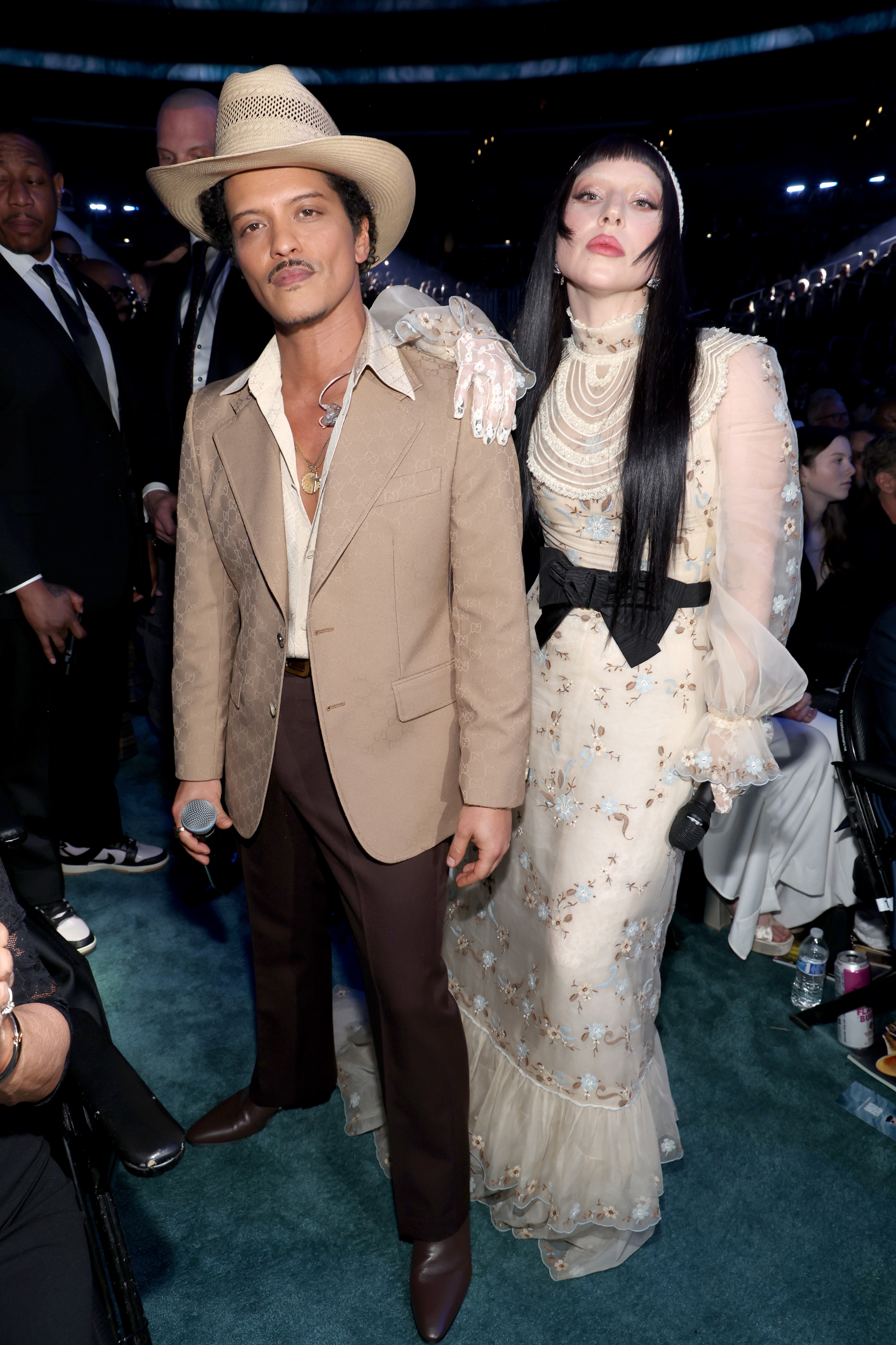 Two radical   astatine  an event, 1  successful  a tan suit   and hat, the different   successful  a agelong  floral formal  with sheer sleeves and melodramatic  makeup