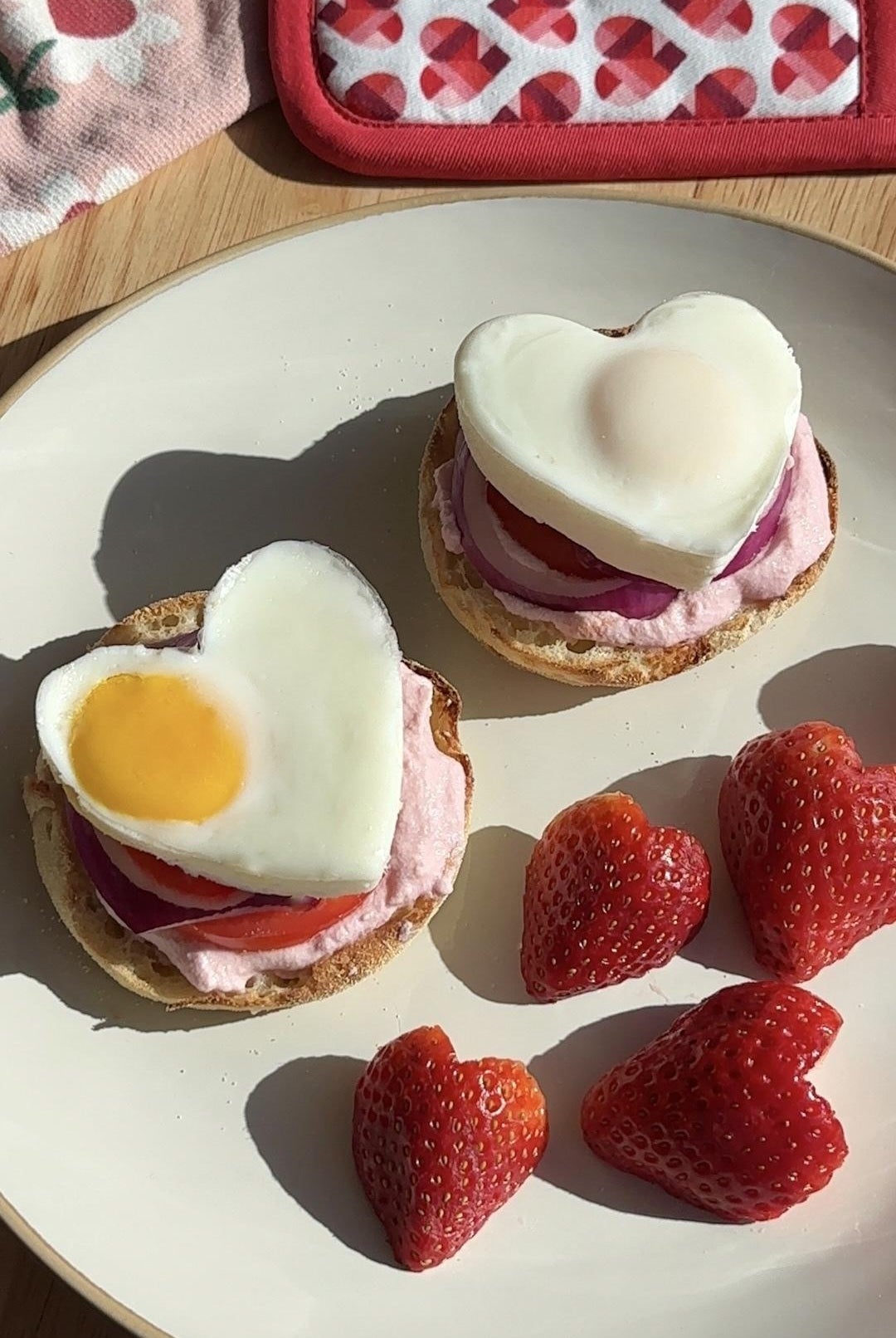 Plate with heart-shaped eggs on toast with strawberries, set on a wooden table with heart-patterned napkin and potholder in the background