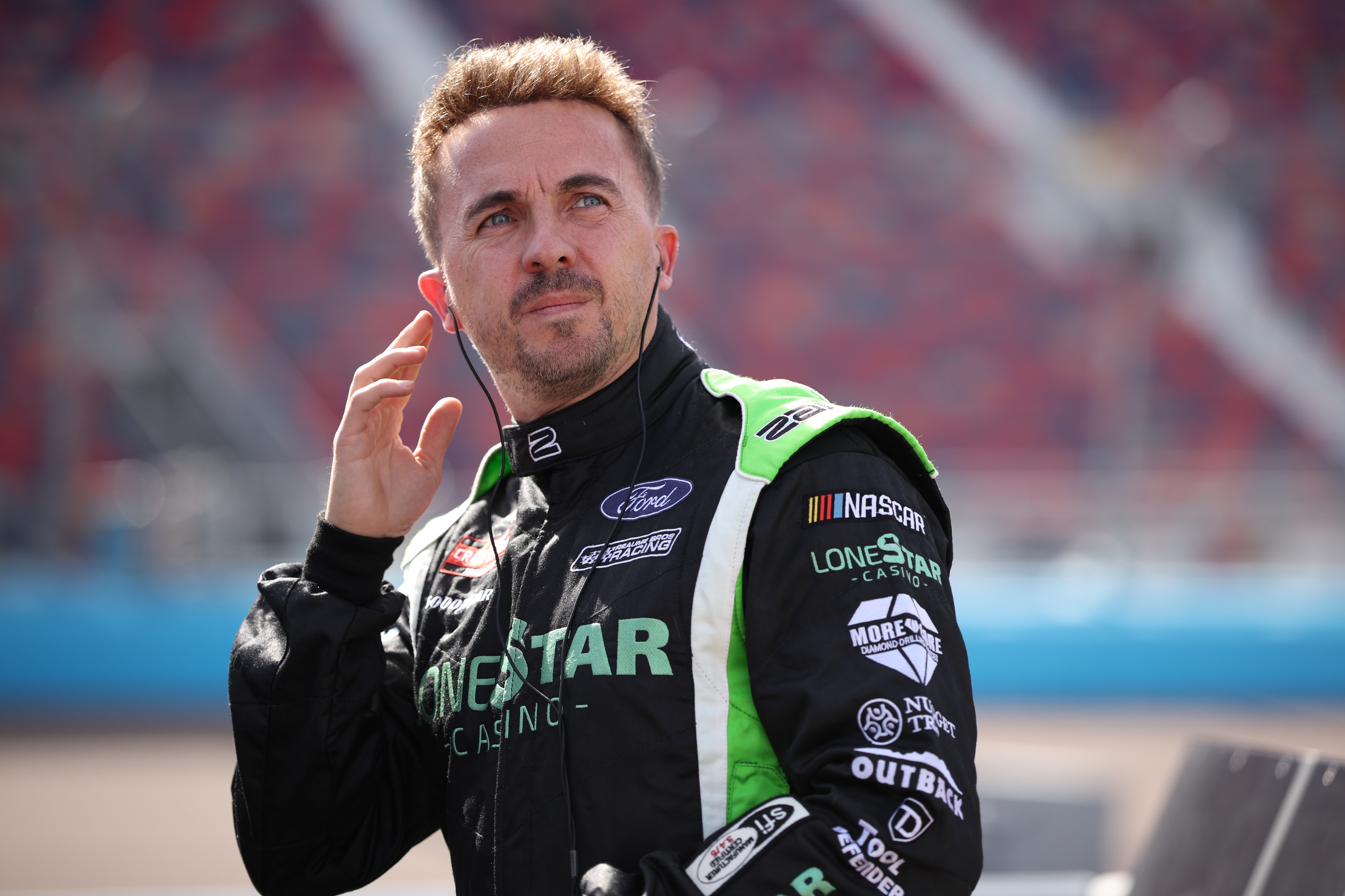 A antheral   successful  a racing suit   adjusts his earpiece astatine  a motorsport event. His suit   features sponsor logos, reflecting a nonrecreational  racing setting