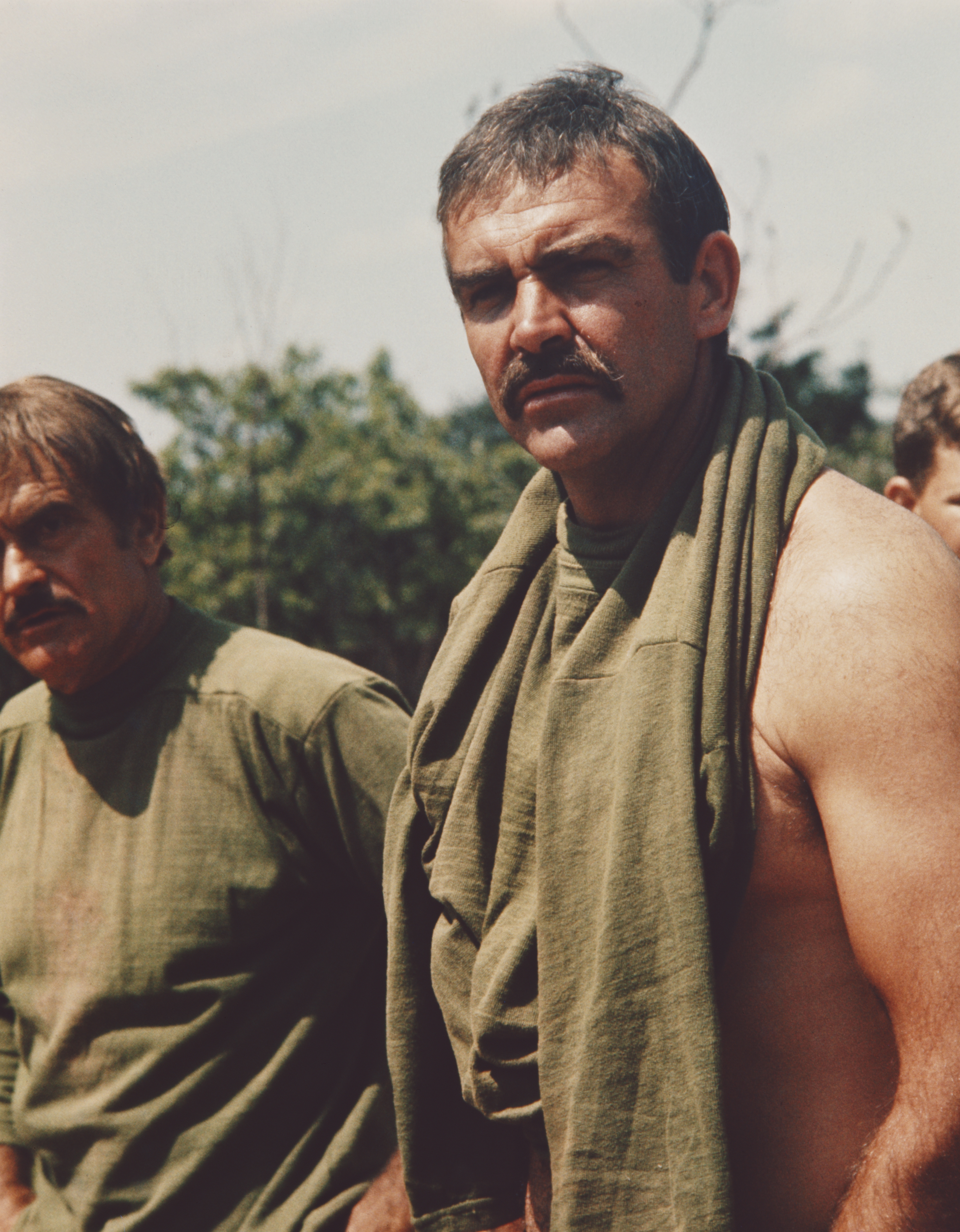 Two men outdoors; 1  has a towel draped implicit    his shoulder. Both deterioration  casual shirts. They look  serious, successful  a earthy  setting