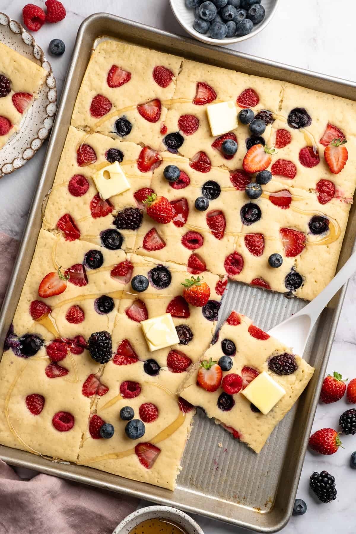 Sheet pan pancakes topped with strawberries, blueberries, blackberries, and sliced butter. A slice is being served