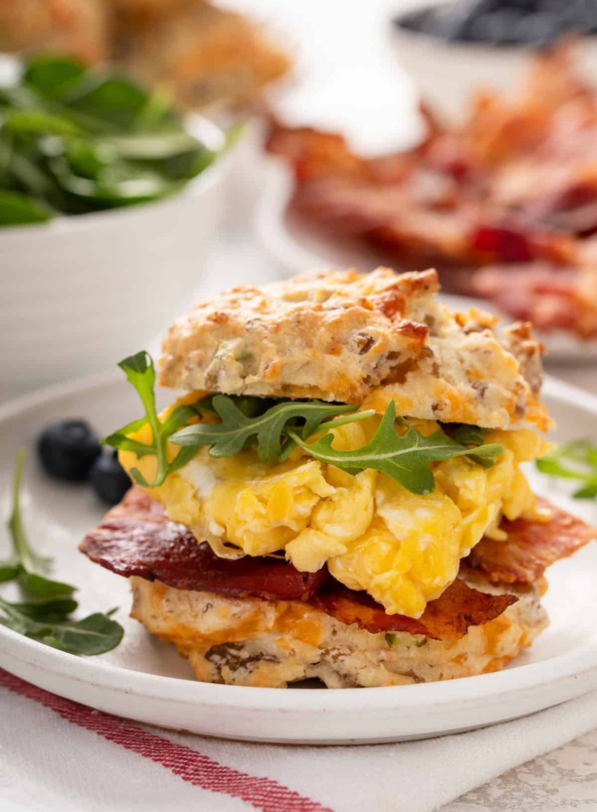 Breakfast biscuit sandwich with scrambled eggs, bacon, and arugula on a plate, with bowls of salad and blueberries in the background