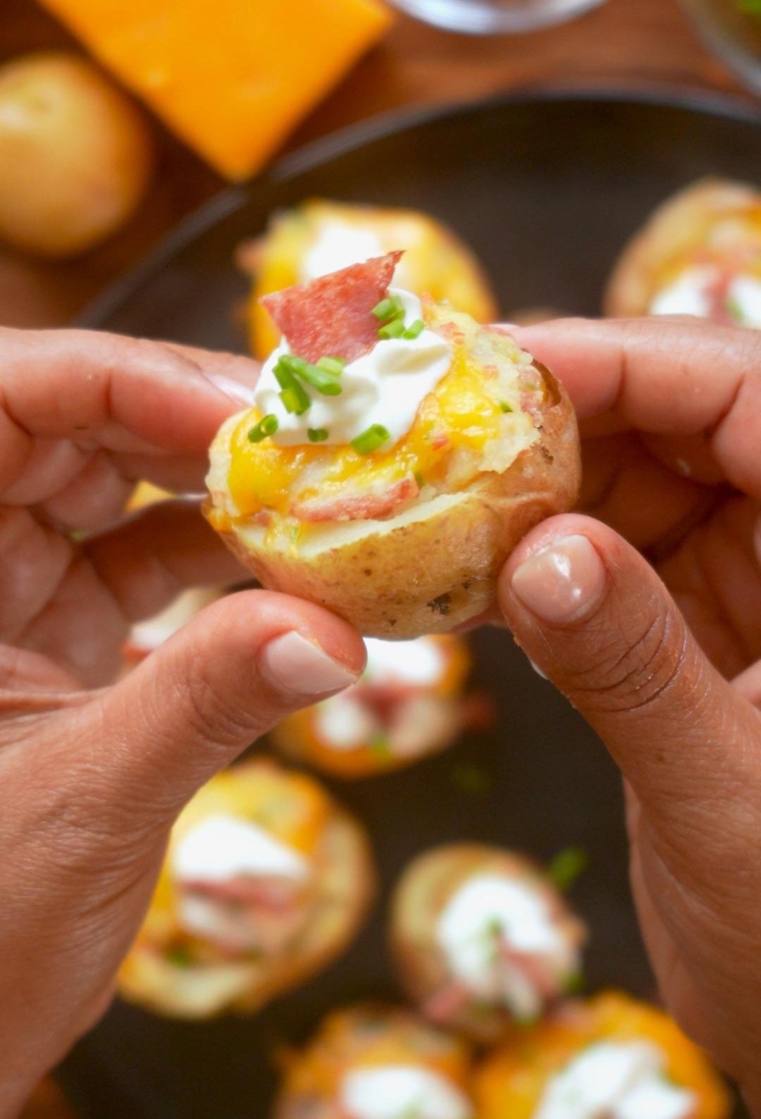 Close-up of hands holding loaded potato bite with cheese, sour cream, and bacon. Several more bites are arranged on a platter in the background