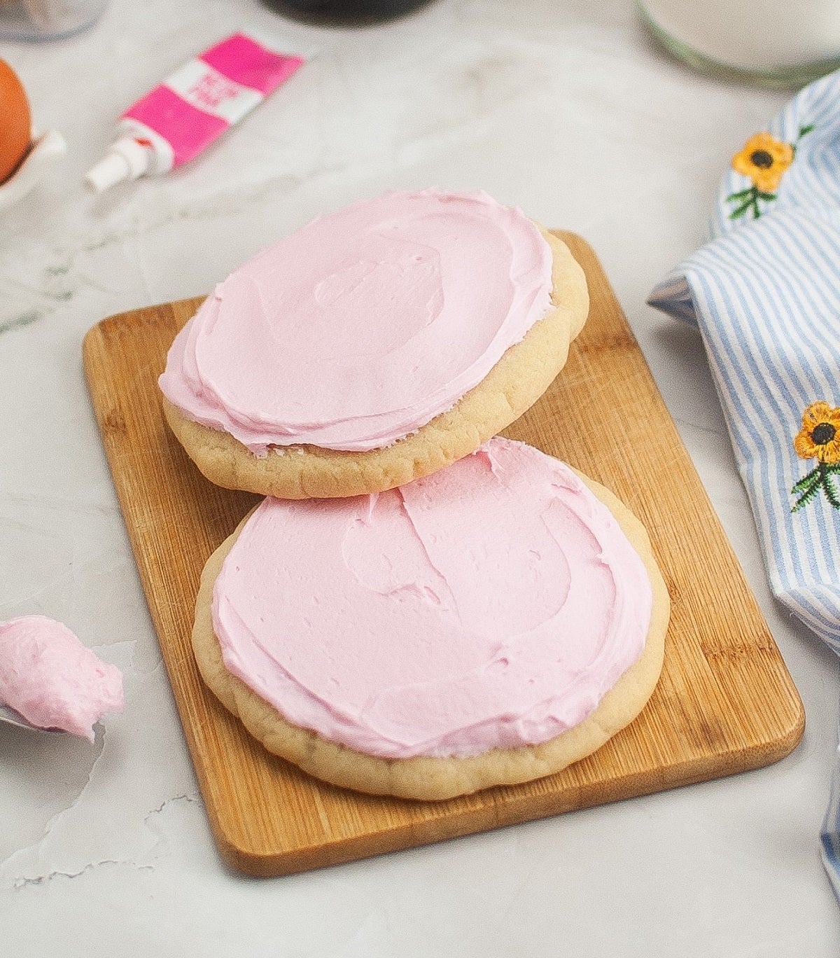 Two sugar cookies with pink frosting on a wooden board. Baking supplies and a striped cloth with floral embroidery are nearby