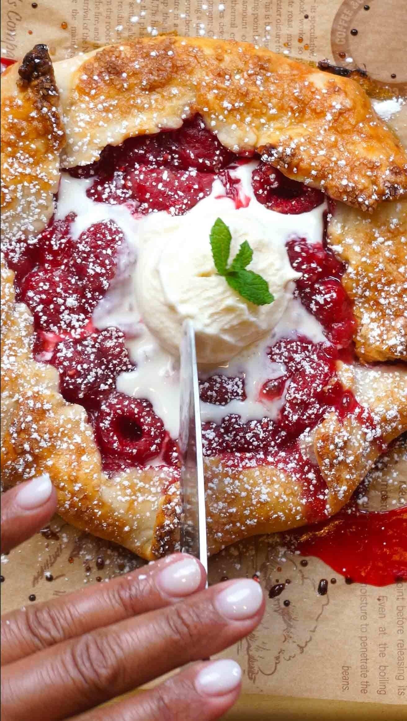 Close-up of a fruit galette topped with a dollop of ice cream and powdered sugar. A hand is slicing into it with a knife