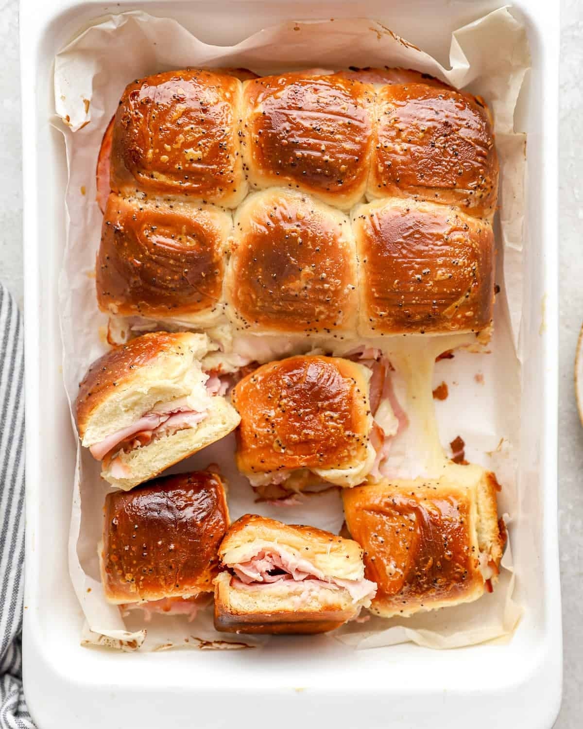 Baked slider sandwiches with ham and cheese, topped with a seasoned glaze, arranged in a rectangular dish on parchment paper