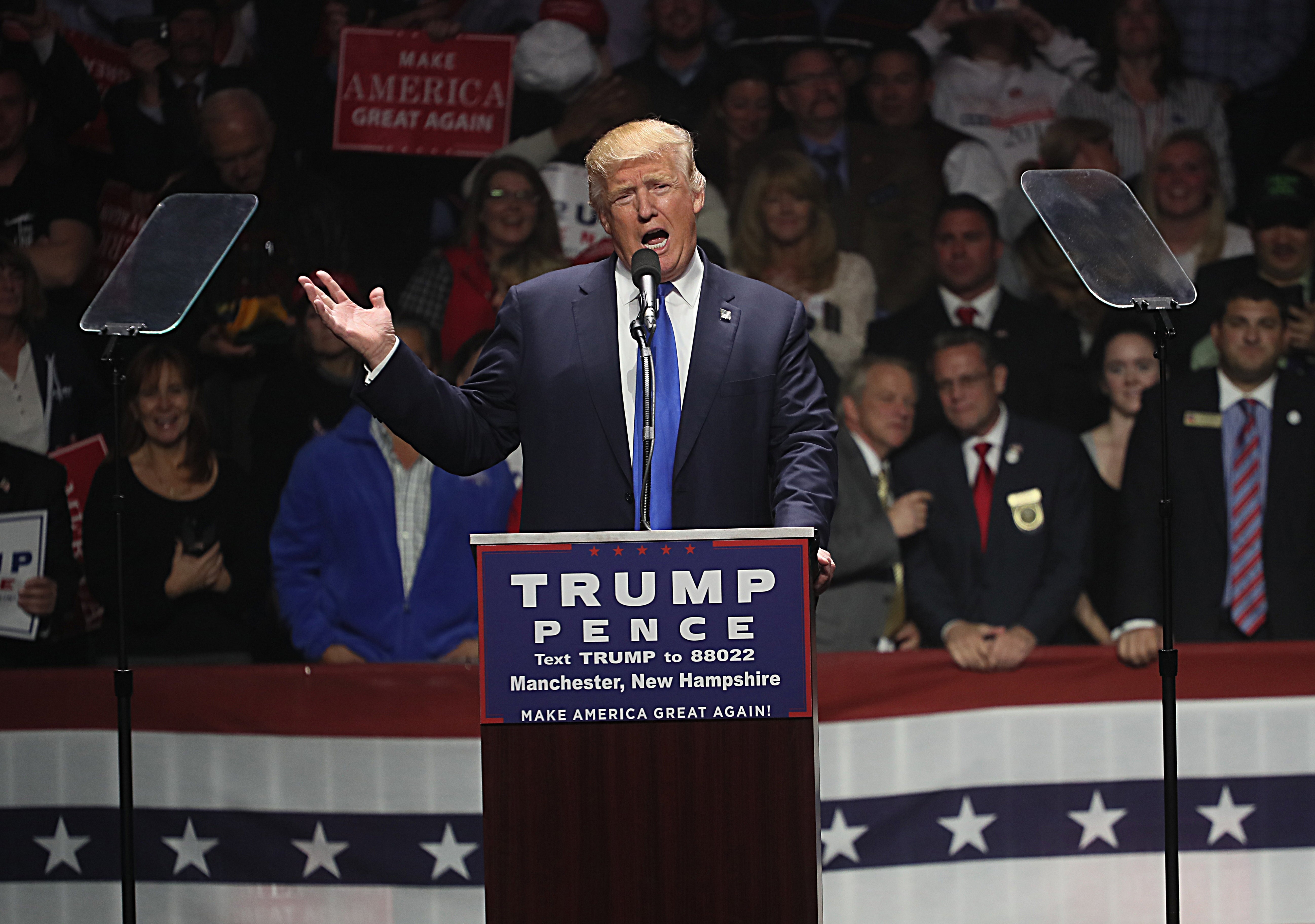 En mann i dress snakker lidenskapelig på et politisk møte, med folk og kampanjeskilt i bakgrunnen