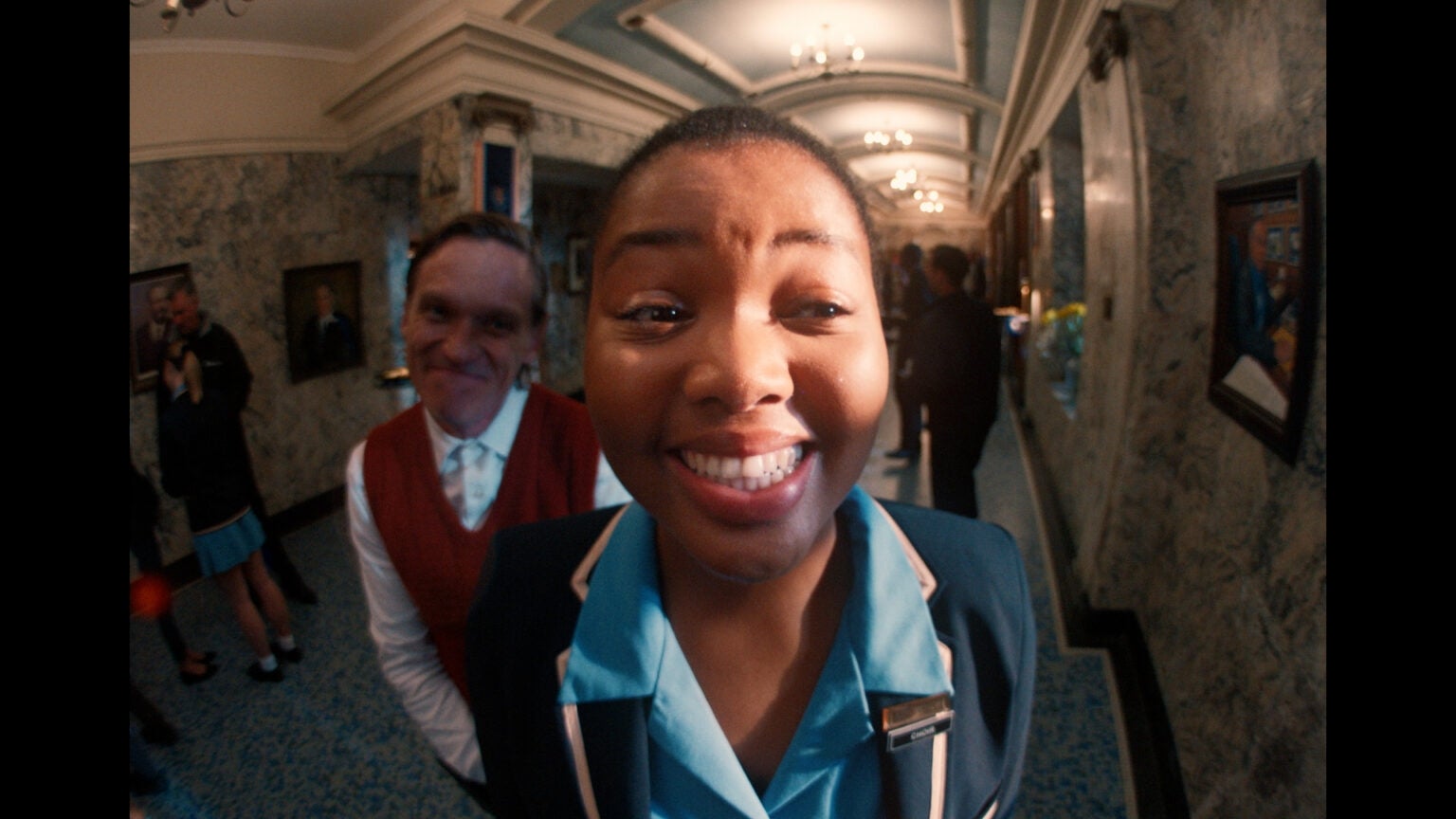 A woman in a blue uniform smiles at the camera in a busy hallway, with a man in a red vest standing behind her