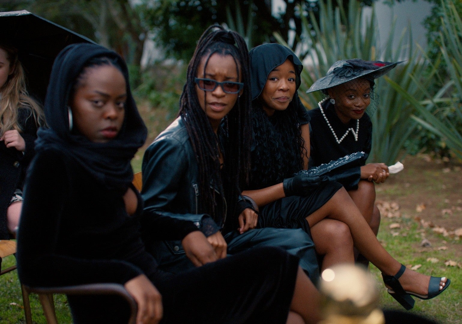 Four people sit outdoors, dressed in stylish black attire and hats, suggesting a somber or formal event