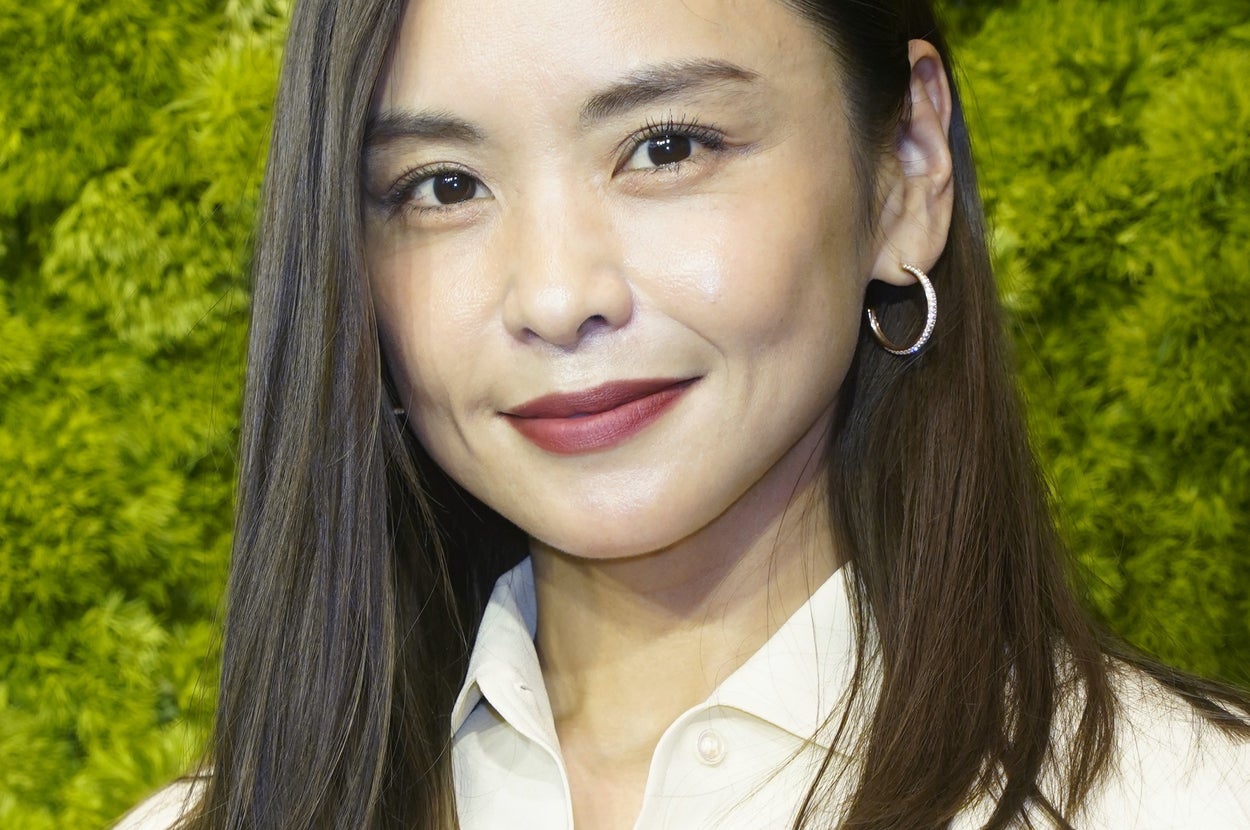 女性が笑顔でカメラを見つめている。背景は緑の植物。彼女はシンプルな白いトップスを着ている。