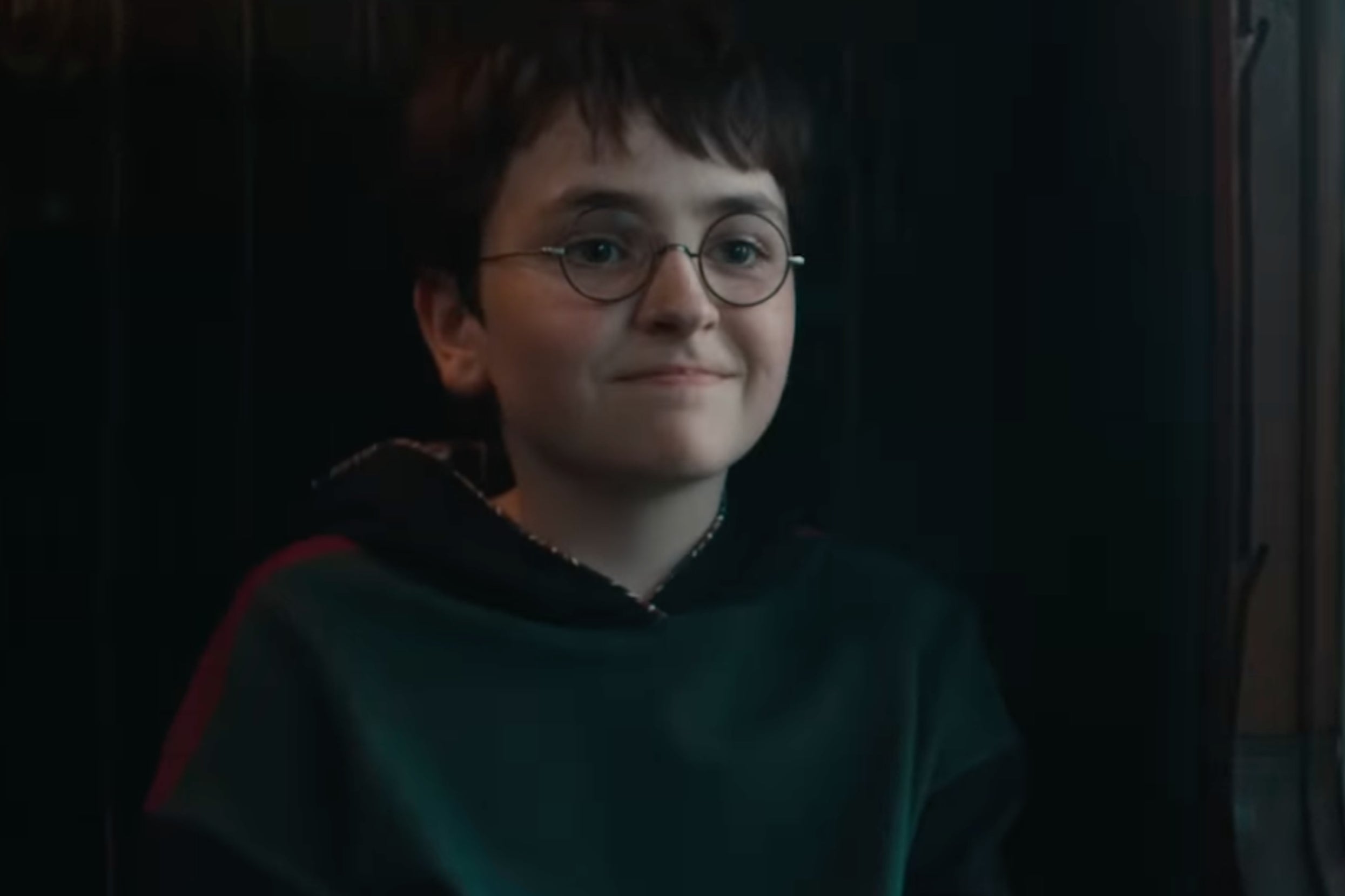 A young person with short hair and glasses smiles, wearing a dark robe-style top, seated indoors