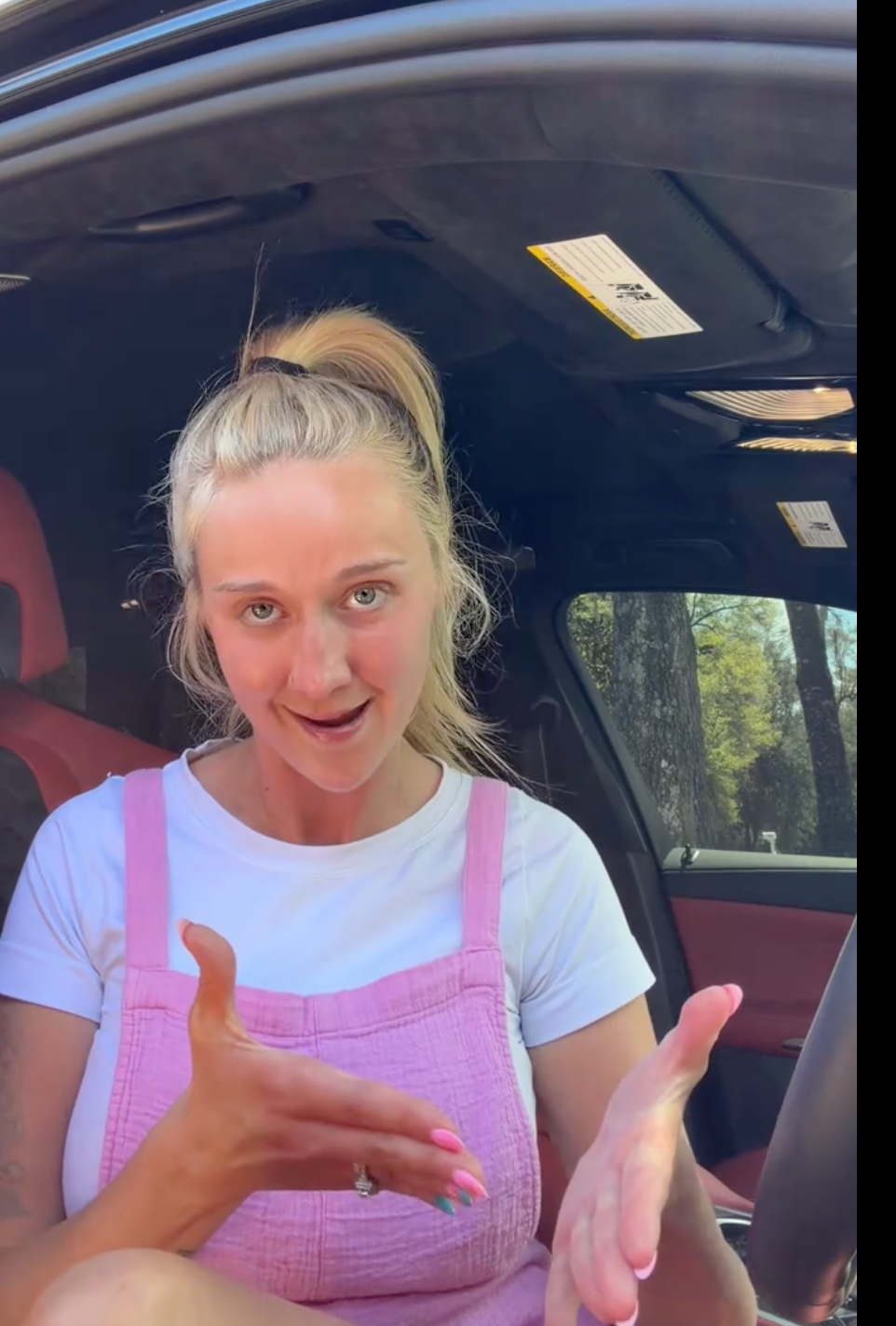 Person sitting successful  a car, wearing a pinkish  wide    formal  and achromatic  t-shirt, gesturing with 1  hand
