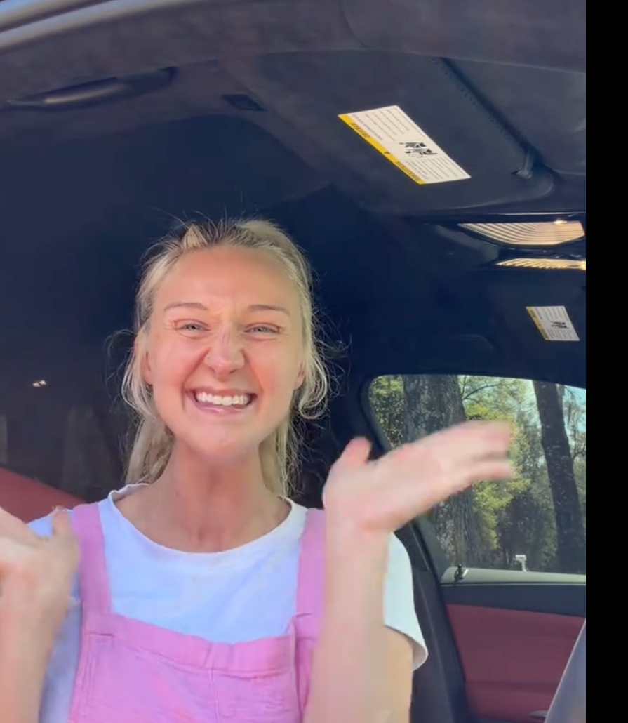 Person smiling wrong  a car, wearing a casual achromatic  t-shirt and pinkish  overalls, gesturing with hands up   successful  excitement