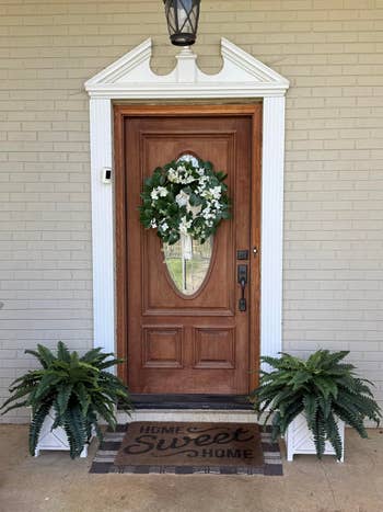 Wooden beforehand doorway with a solid panel, adorned with a floral wreath, flanked by greenish potted ferns, and a