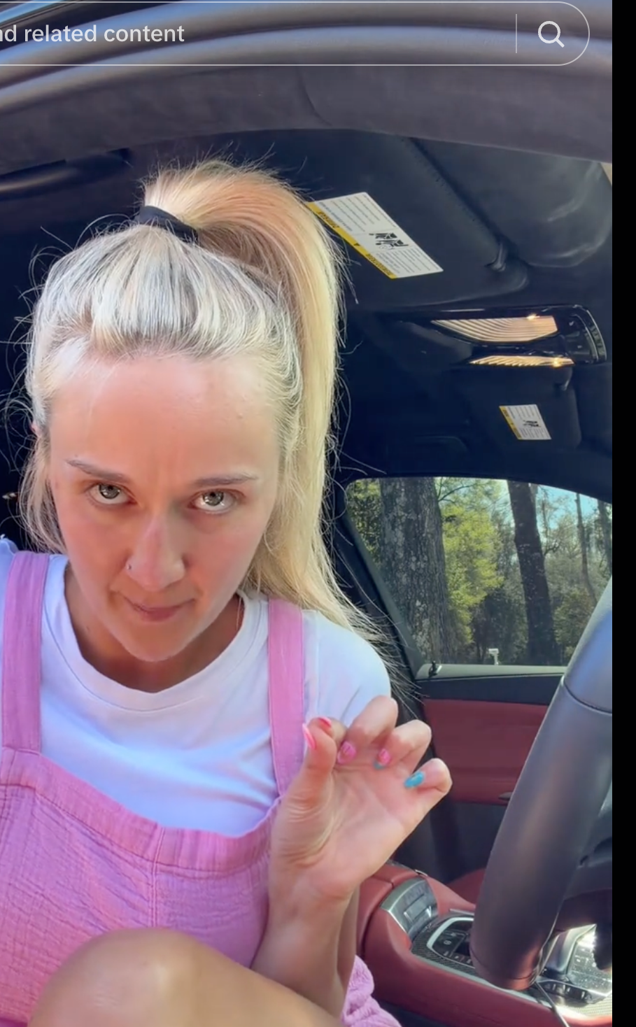 A idiosyncratic   sits successful  a car   wearing a achromatic  garment  nether  pinkish  overalls, with hairsbreadth  successful  a precocious   ponytail, gesturing towards the camera