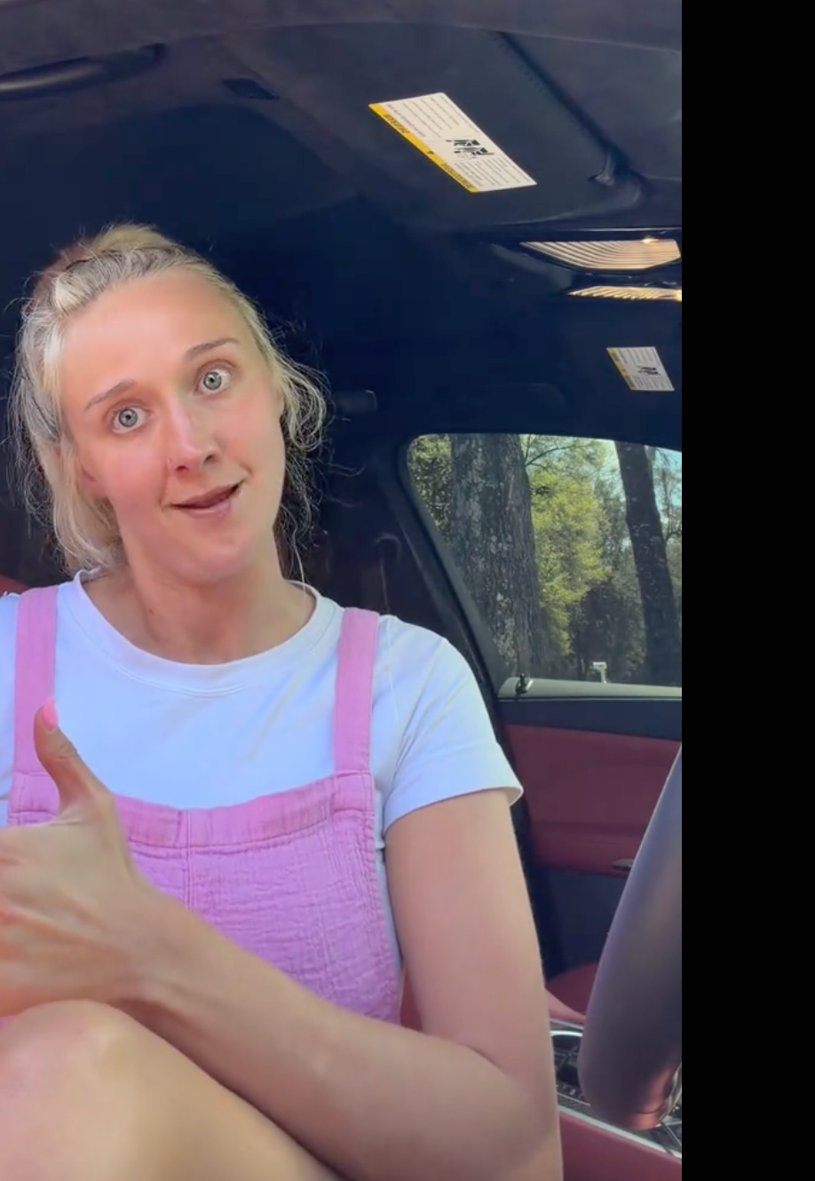 Person sitting successful  a car   seat, wearing a casual light-colored apical  with pinkish  overalls, looking astatine  the camera with a playful expression
