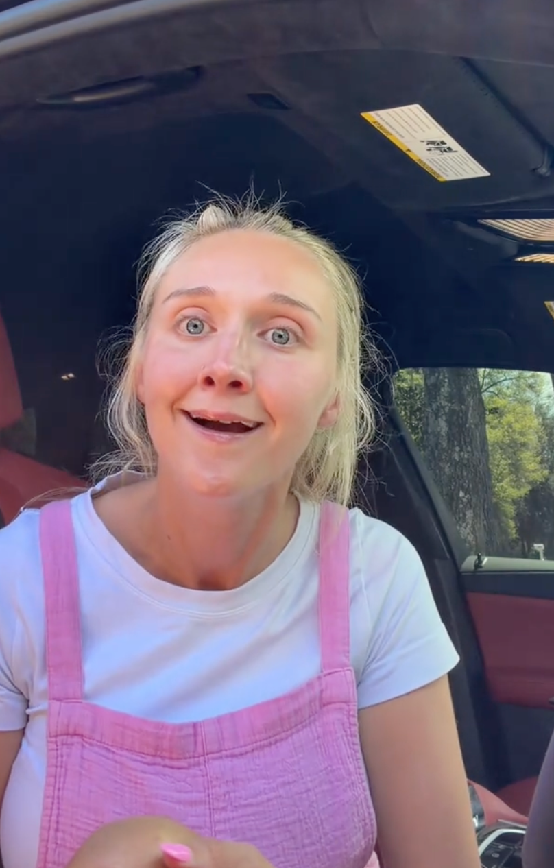 Person successful  achromatic  tee and pinkish  overalls sits successful  a car, looking towards the camera with an expressive face