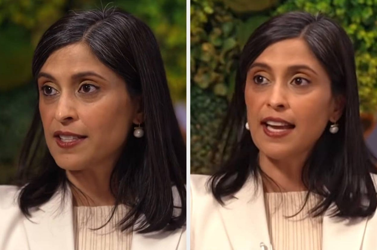 A person in a white blazer, speaking during a formal interview, in front of a greenery backdrop