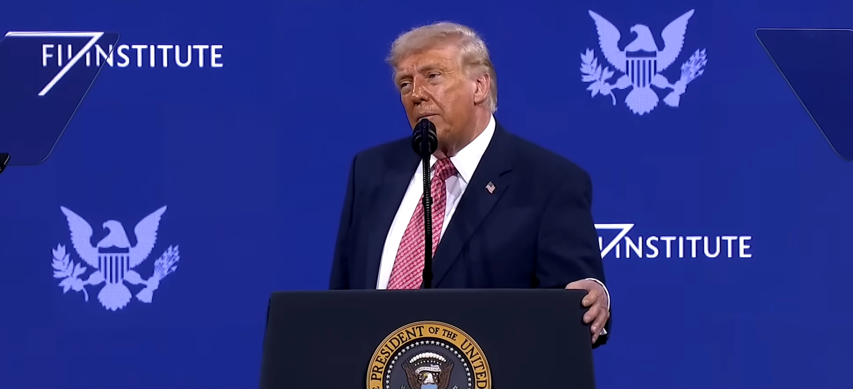 A idiosyncratic speaking astatine a podium with a seal, against a bluish backdrop with the "America First Policy Institute" logo