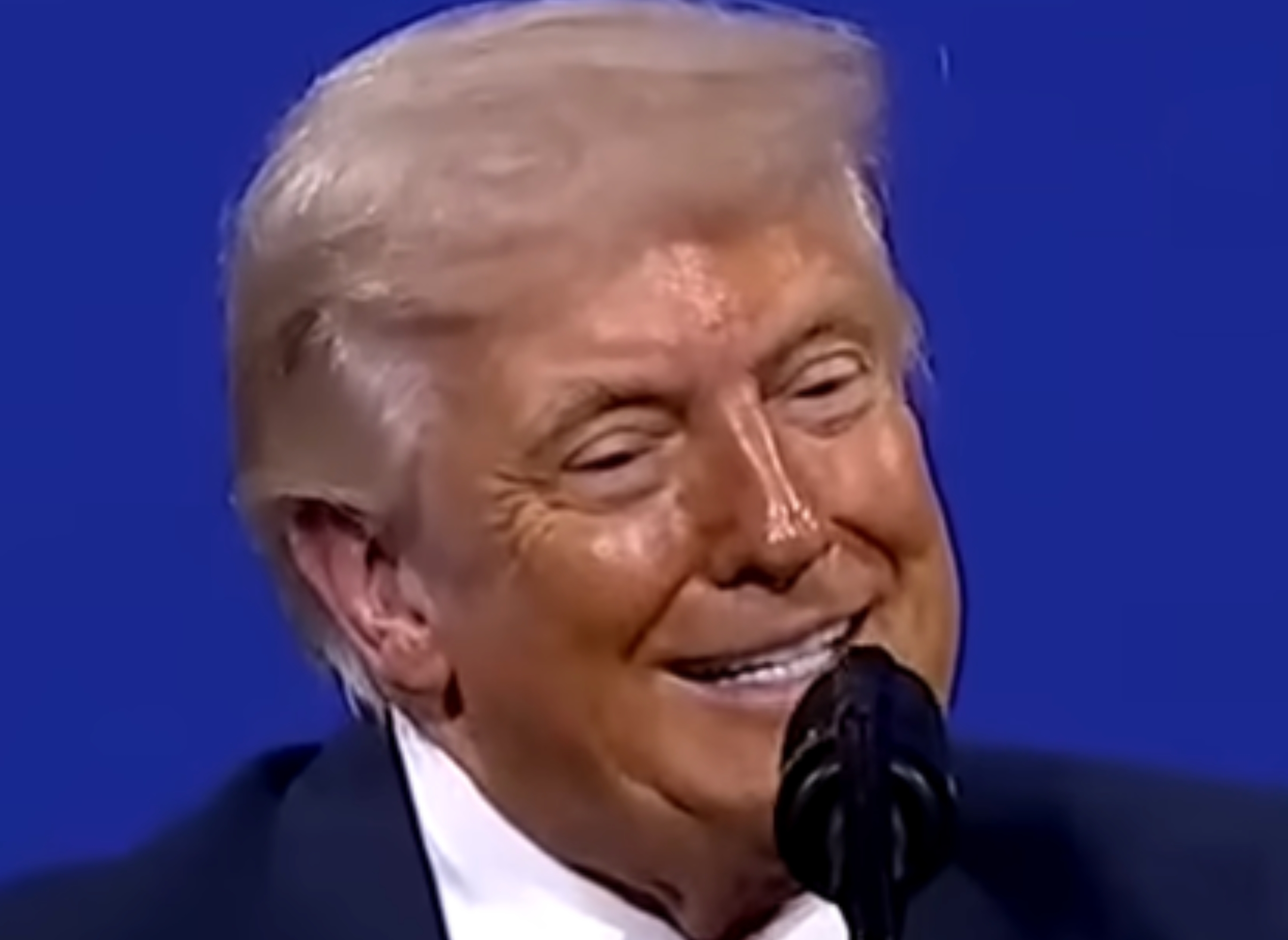 A idiosyncratic successful a suit and necktie speaks astatine a podium with a seal, successful beforehand of a bluish backdrop with abstract logos and text