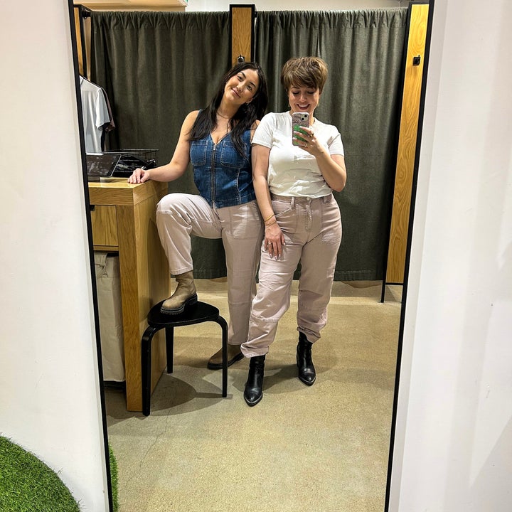 Two people in casual outfits pose in a clothing store mirror. One leans on a stool; the other takes a selfie