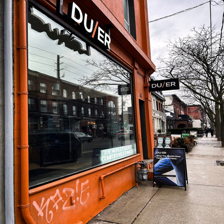 Storefront of DUER with sidewalk sign highlighting performance denim. Street scene includes trees and neighboring shops