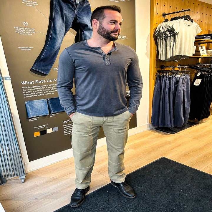 A person models casual clothing in a retail store, wearing a long-sleeve henley shirt and light-colored pants, next to a rack of jeans and tops