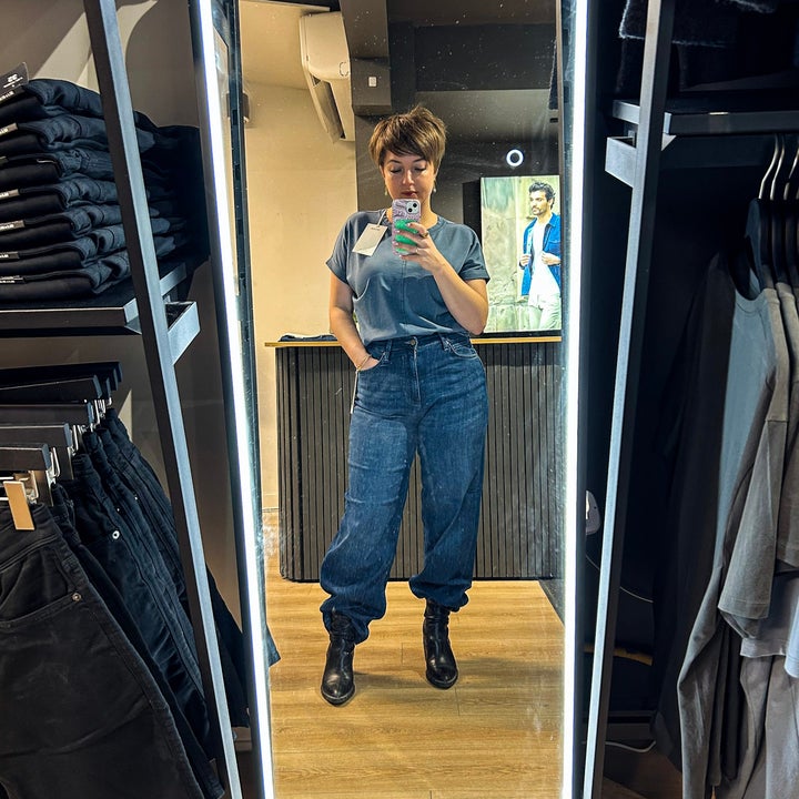Person wearing a casual top and loose-fitting jeans, taking a mirror selfie in a clothing store