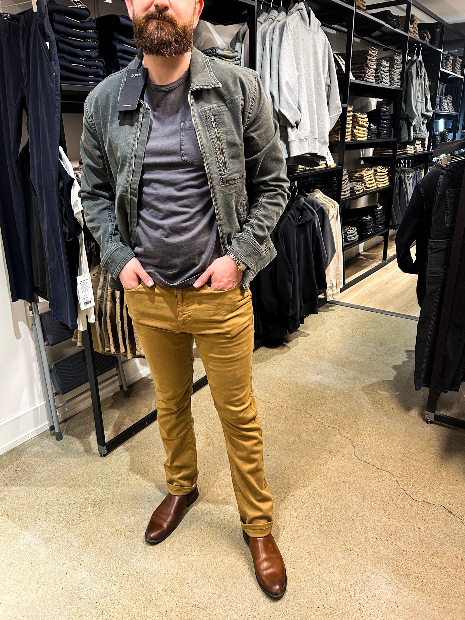  a rugged jacket, acheronian  shirt, mustard pants, and brownish  boots, surrounded by racks of folded clothes