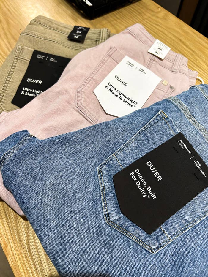 Three pairs of DUER jeans successful  beige, pink, and bluish  are displayed connected  a woody  table, highlighting tags emphasizing comfortableness  and design