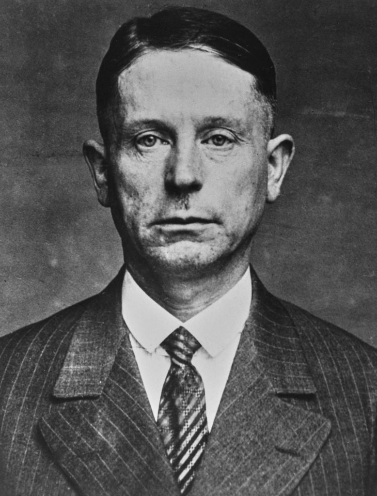 A historical black and white portrait of a man in a pinstripe suit and tie, looking directly at the camera