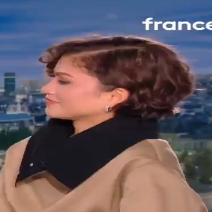 Person with short hair in a studio, wearing a stylish outfit with a large collar, cityscape in the background. News channel logo and time shown