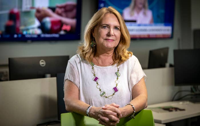 A pistillate   sits successful  an bureau   setting, wearing a casual blouse and a beaded necklace. Monitors are disposable   successful  the background