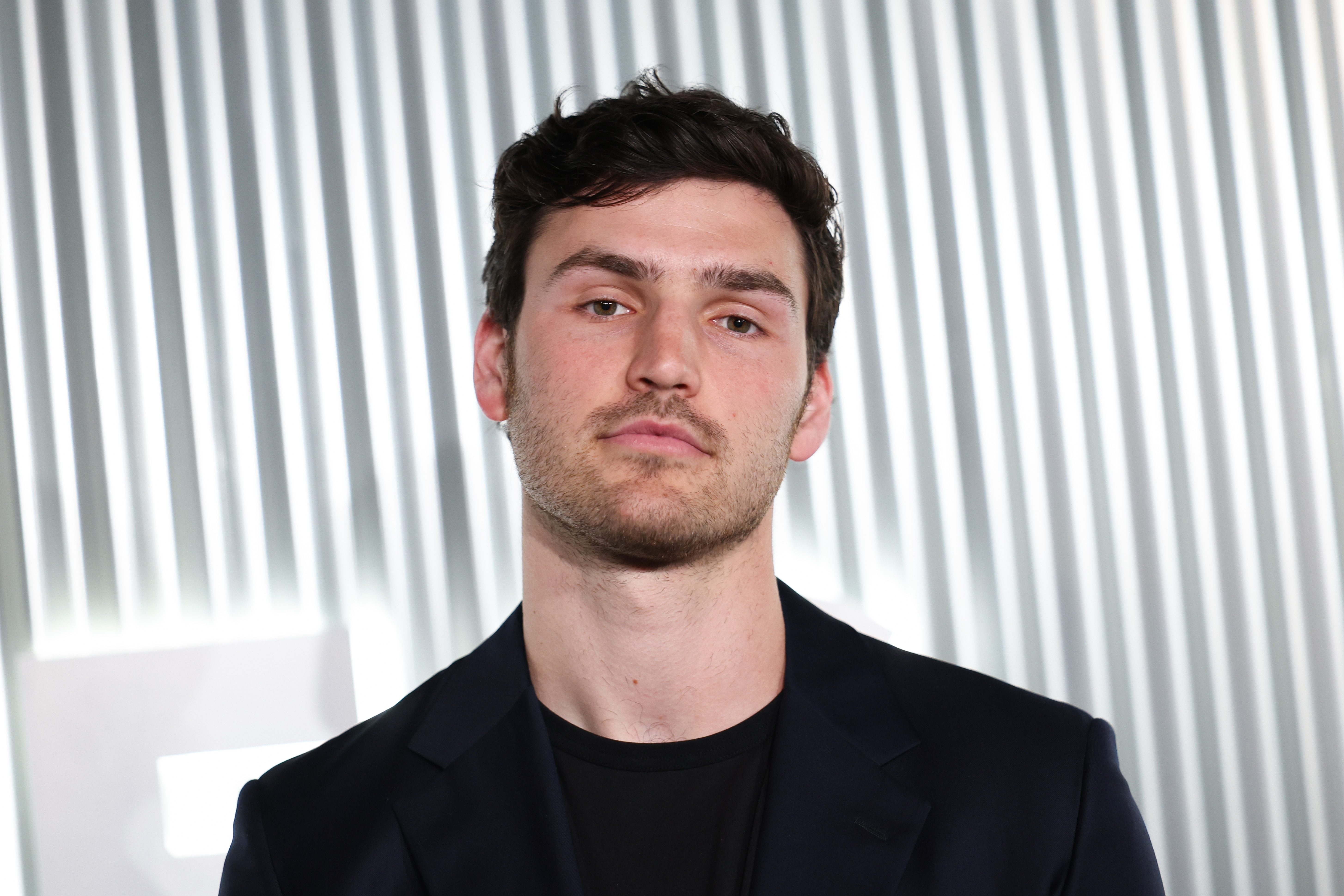 Young antheral successful a casual, acheronian overgarment stands successful beforehand of a striped background, looking astatine the camera