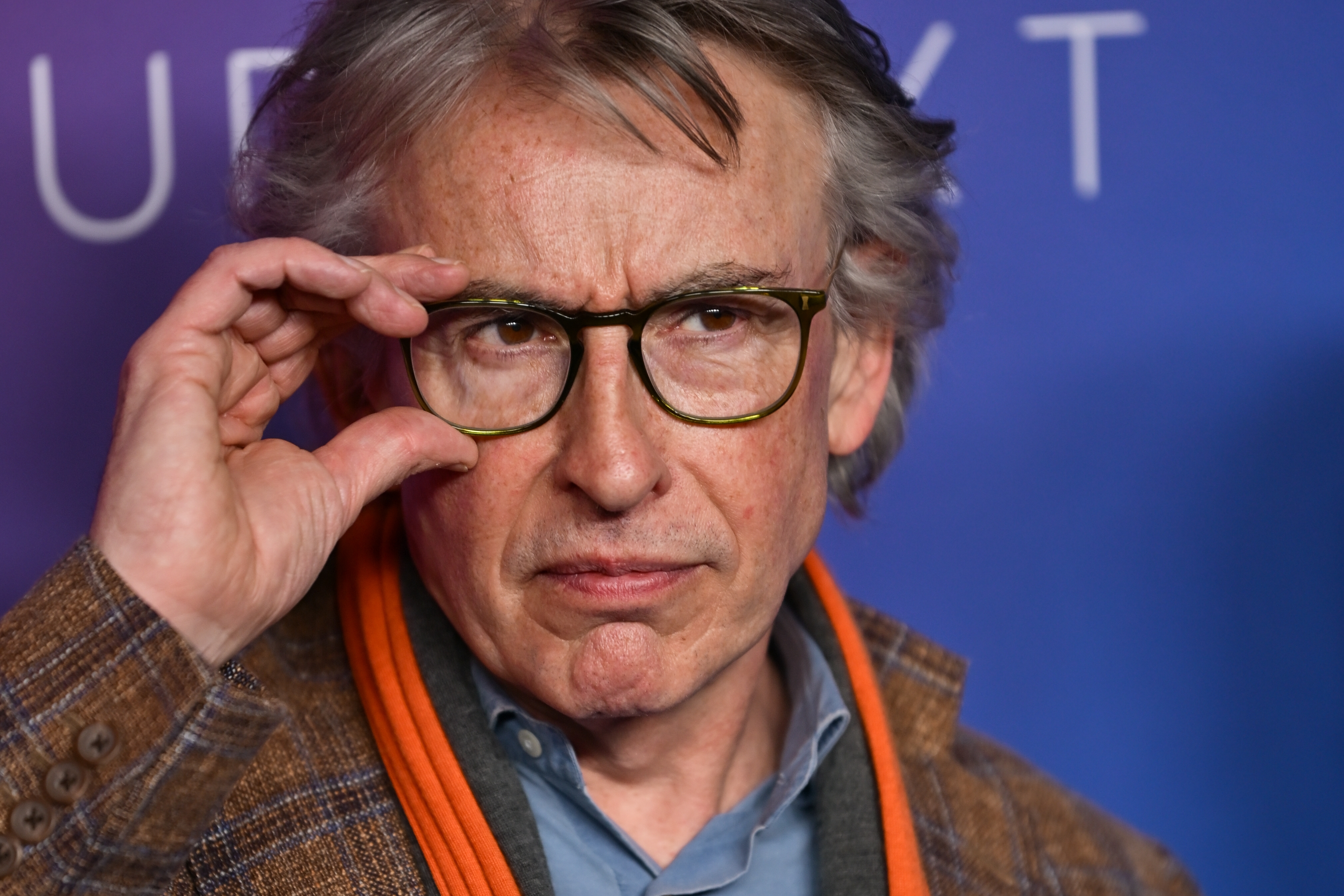 Person with glasses adjusting frames, wearing a plaid overgarment and orangish scarf, against a bluish background