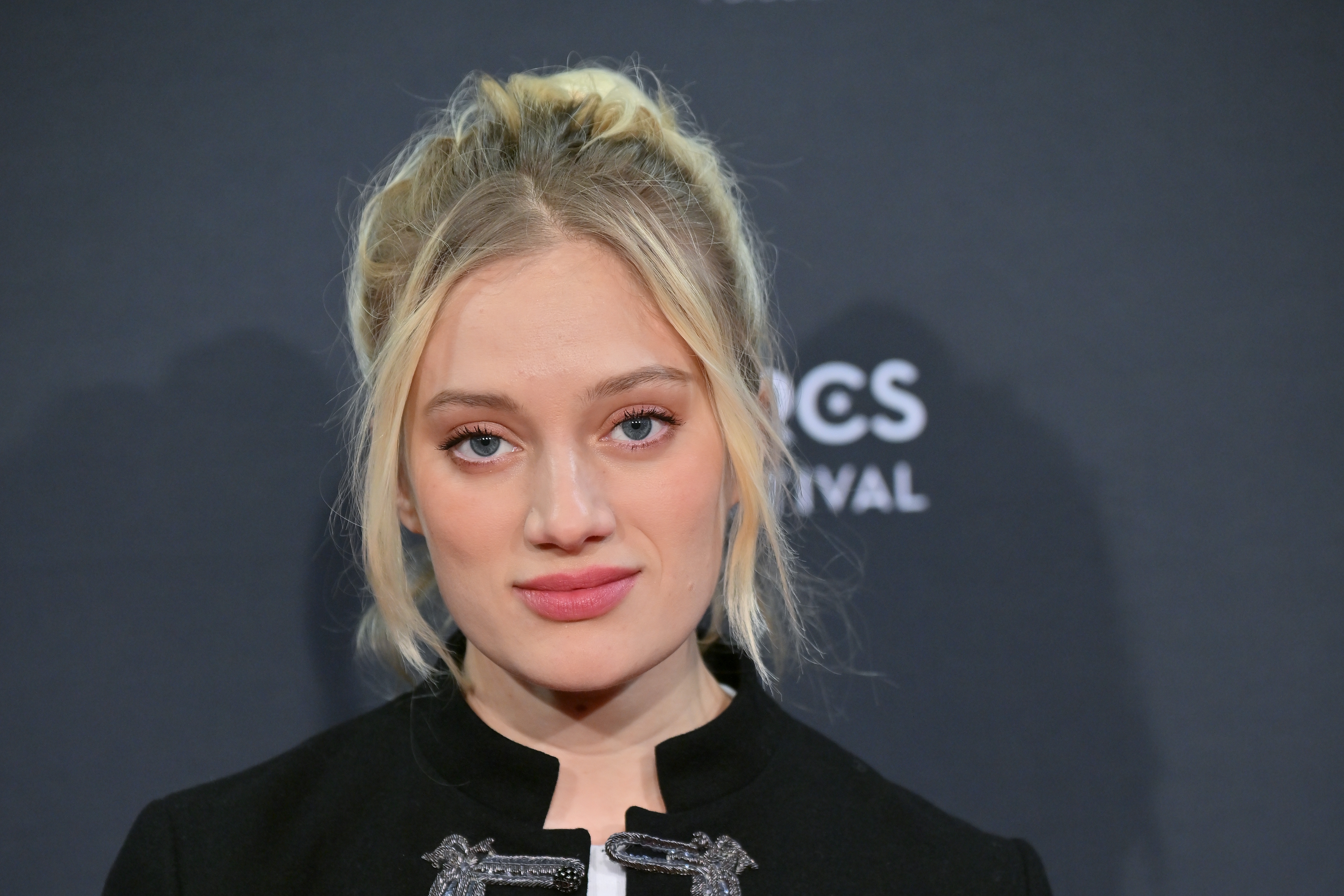 Person with a neutral expression, wearing a ceremonial outfit with decorative accents, stands against a acheronian backdrop with "SPIKES ASIA FESTIVAL" partially visible