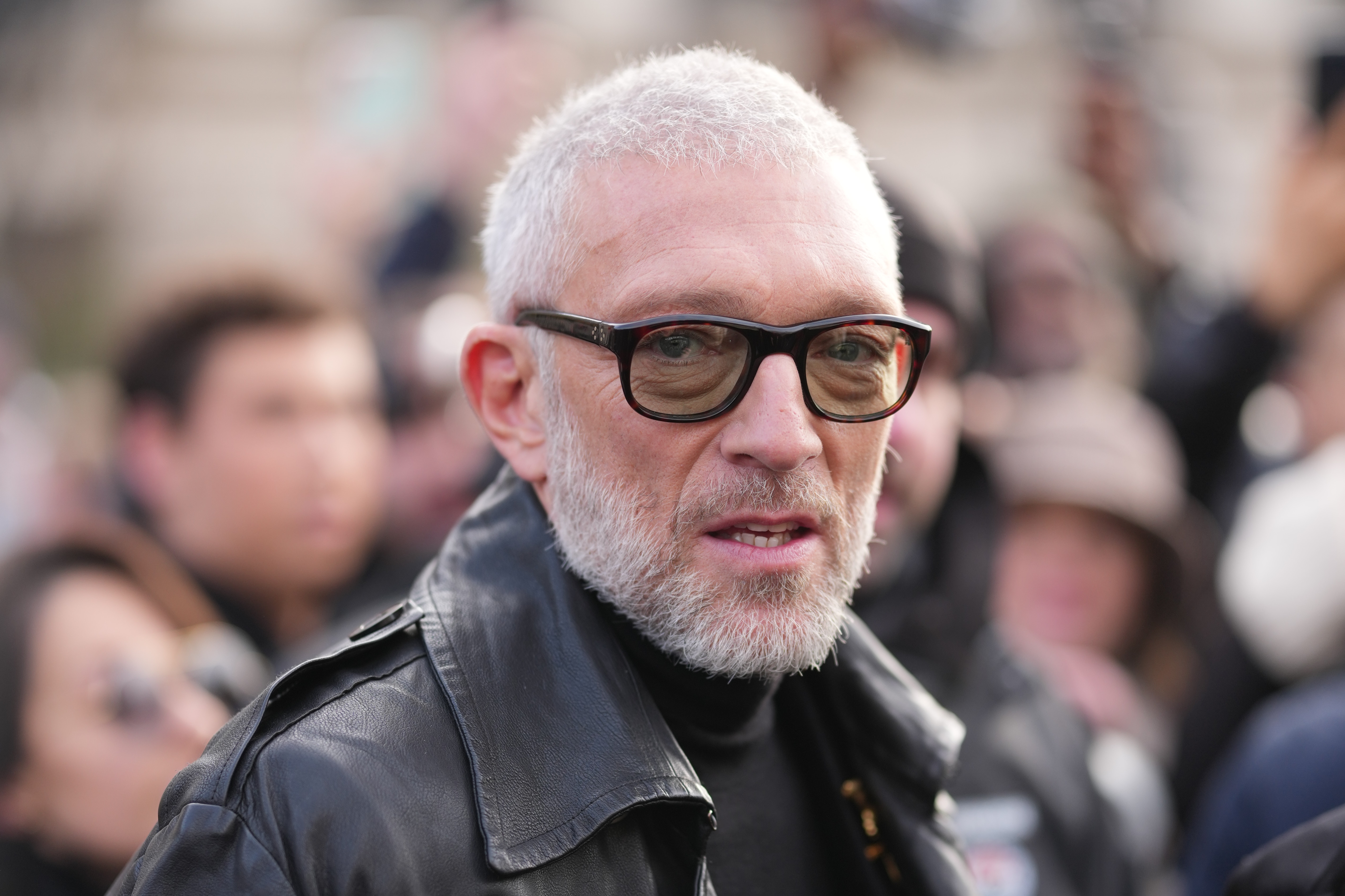 A idiosyncratic with a abbreviated beard and glasses wearing a achromatic leather overgarment looks towards the camera successful a crowded outdoor setting