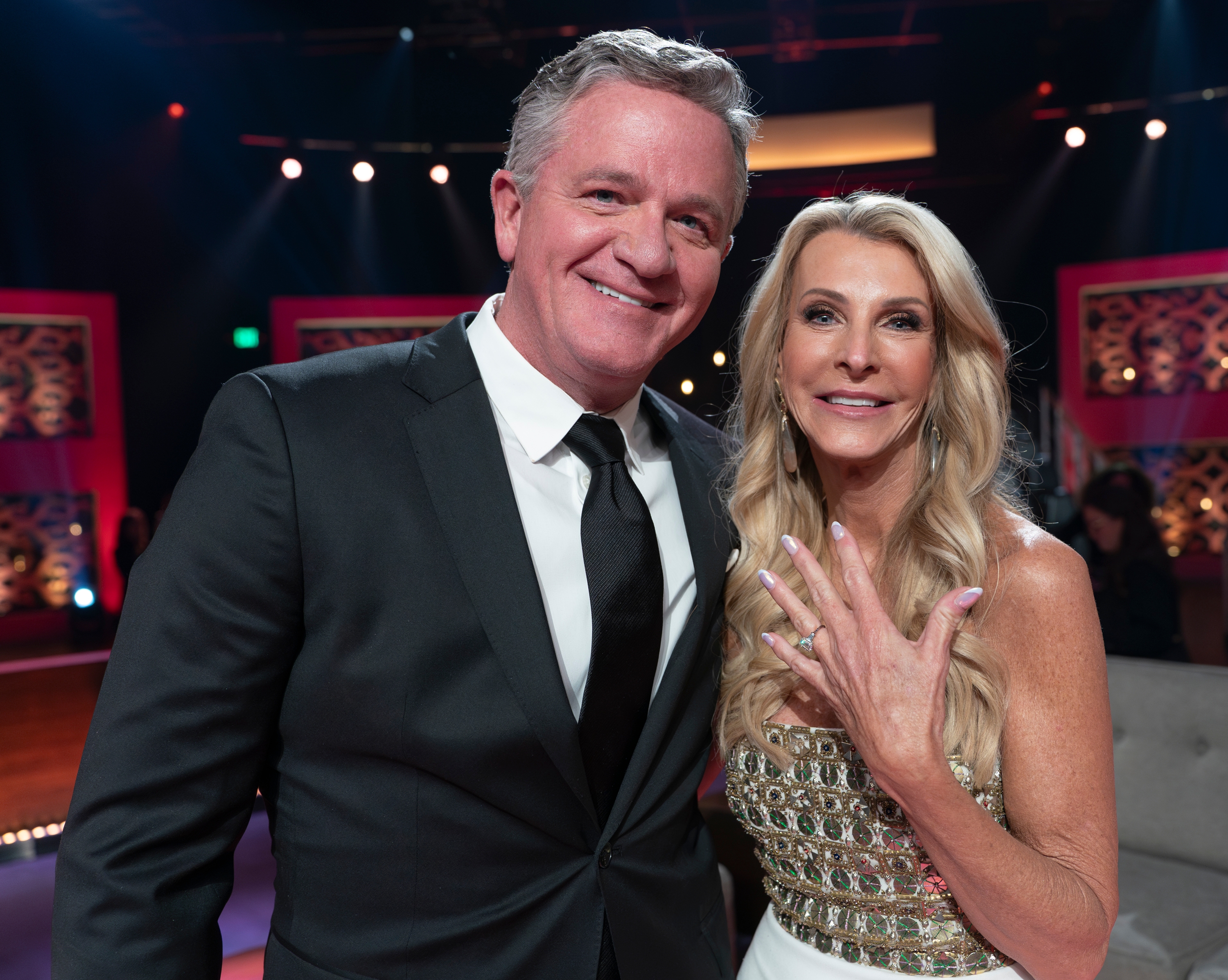 A antheral   successful  a suit   stands beside a pistillate   showing an engagement ring, wearing a formal  with intricate beading, connected  a lit stage