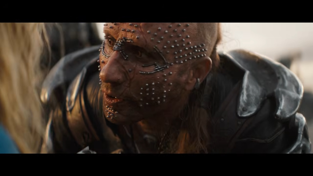 A character with metallic face adornments and armor leans close to another character, in an intense scene from a TV or movie