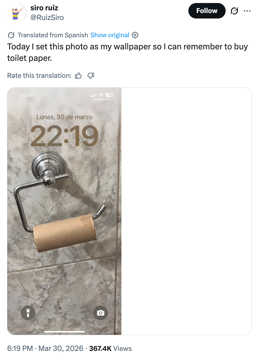 An empty toilet paper roll on a holder, with a phone wallpaper showing the same image and a time and date overlay