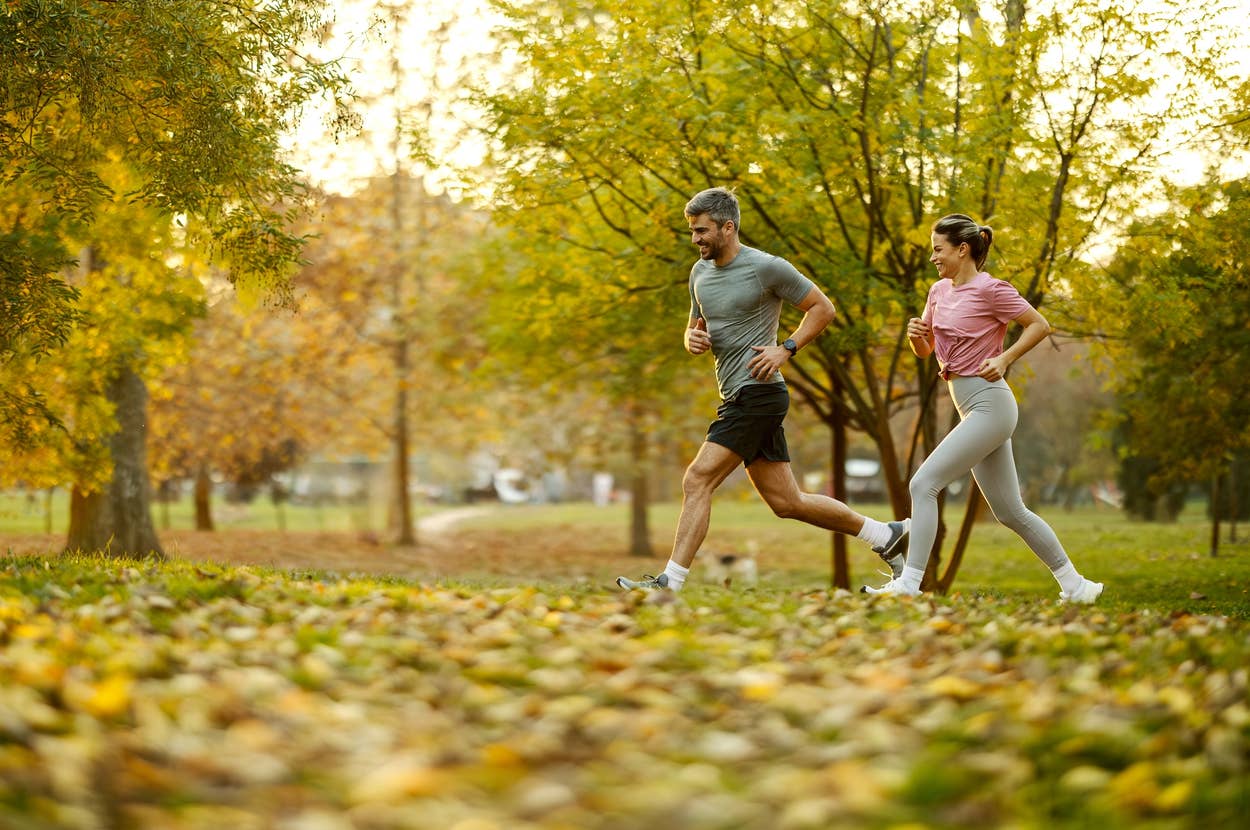Two people jogging in a park surrounded by trees and fallen leaves, enjoying a healthy outdoor activity