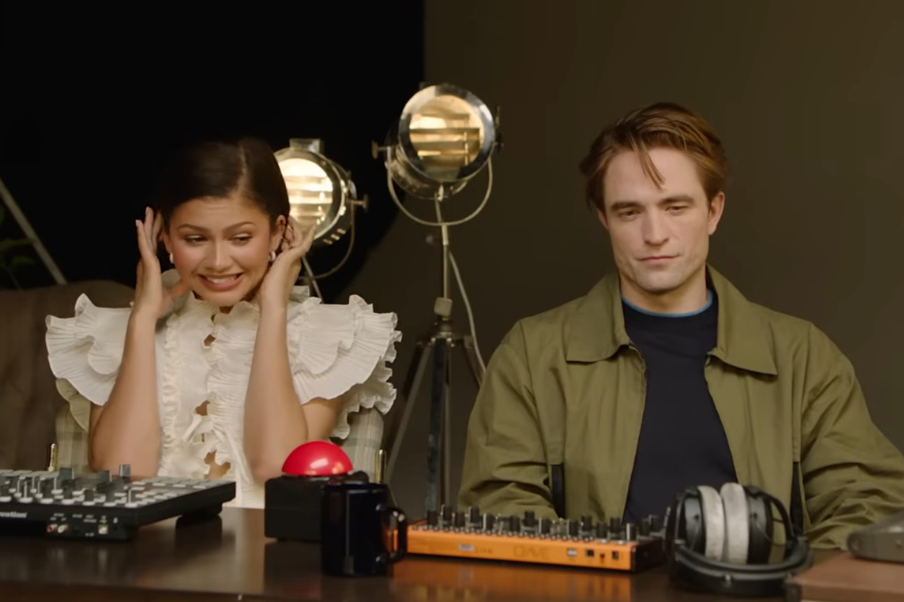 Two people sit at a table with DJ equipment. The person on the left smiles, covering their ears playfully, while the person on the right looks relaxed