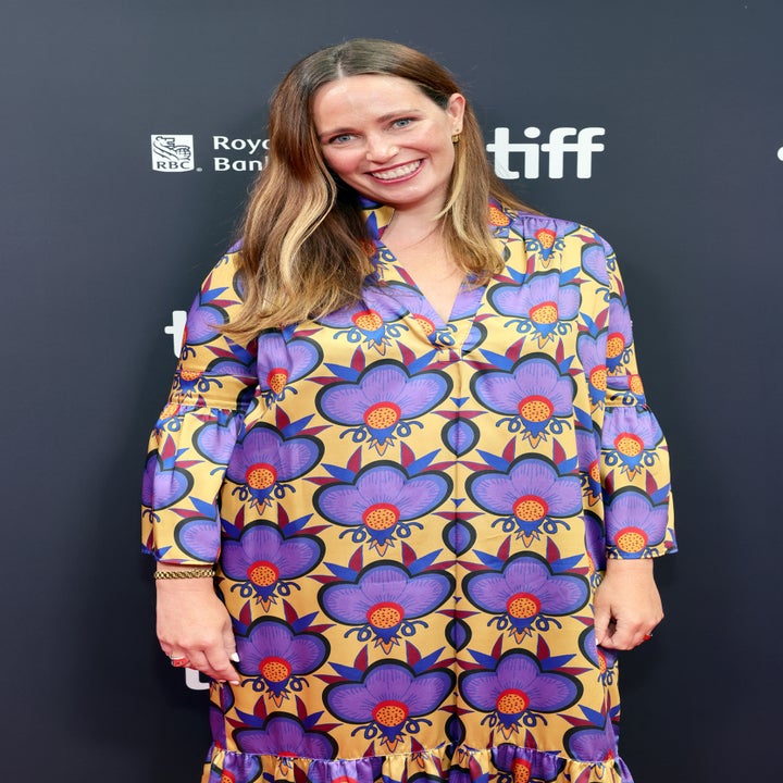 Person smiling in bold floral dress at a film event; standing against a backdrop with sponsor logos