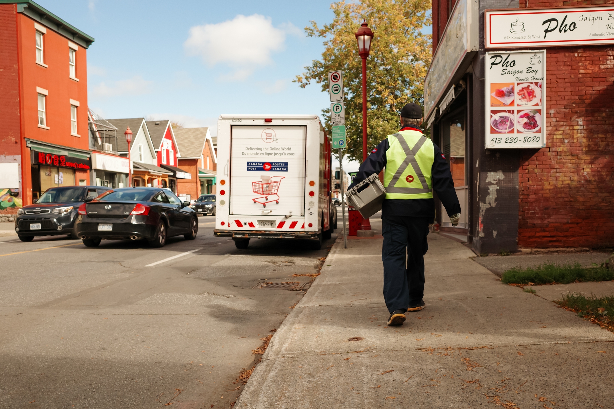 A transportation  idiosyncratic    successful  a reflective vest carries a bundle  connected  a sidewalk beside a thoroughfare  with parked vehicles and adjacent    shops