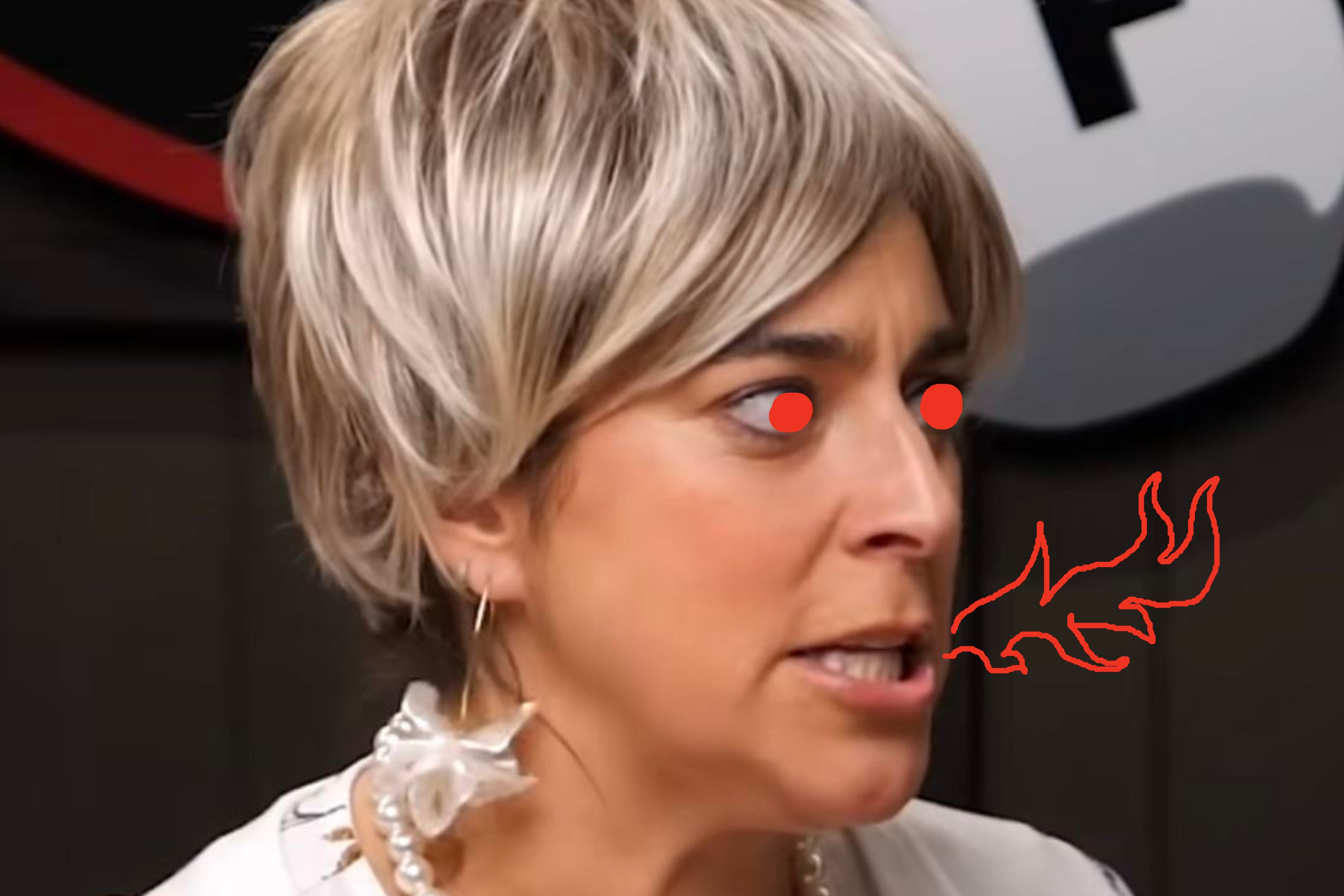 Person with short, light hair looks intense, wearing a pearl necklace and floral earrings. Background is partially obscured