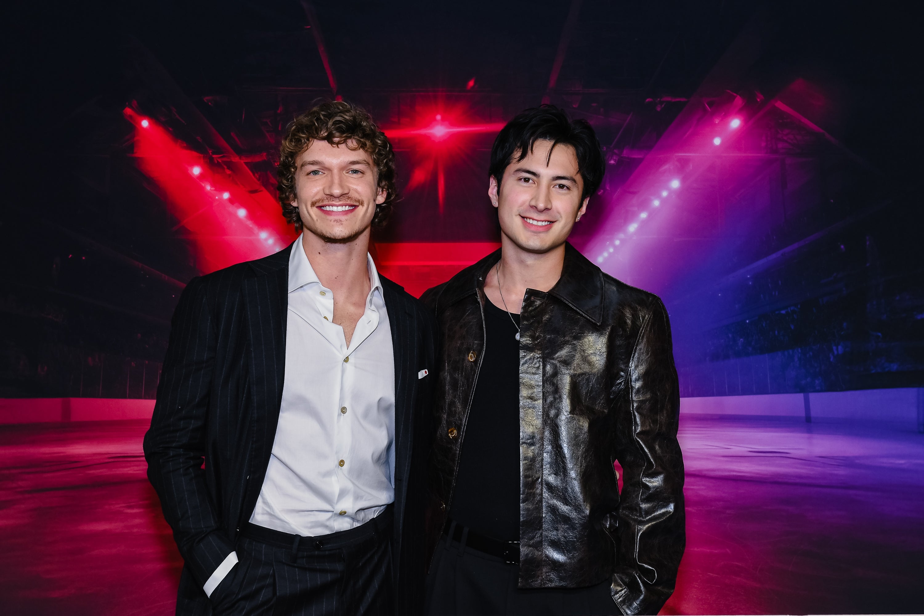 To personer står sammen på en arena med skarpe lys. Begge har på seg stilige jakker, den ene i mønstret dress og den andre i en skinnjakke