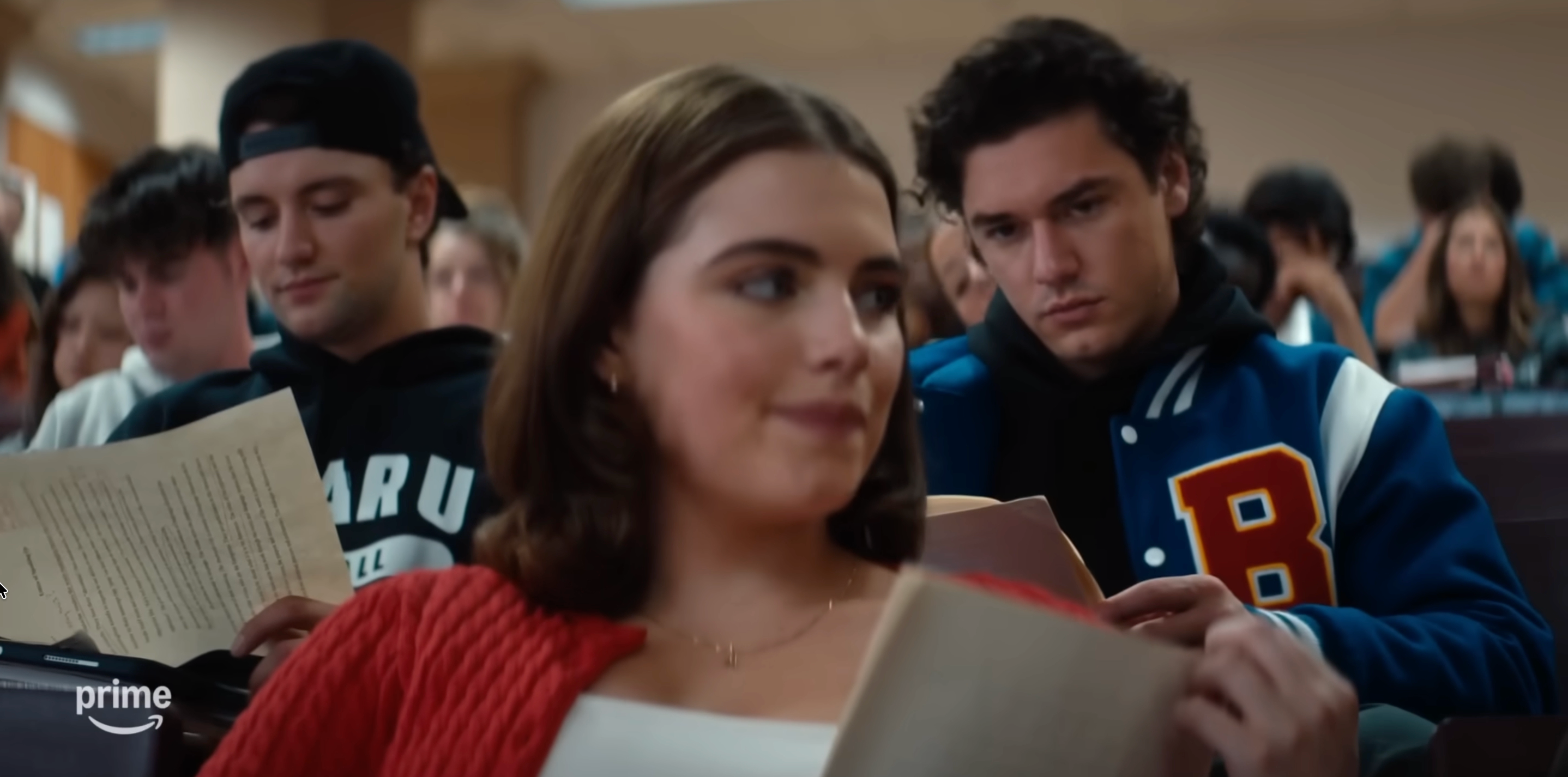 Studenter i en forelesningssal leser artikler. En person i forgrunnen har på seg en rød cardigan, mens en person bak bruker en universitetsjakke