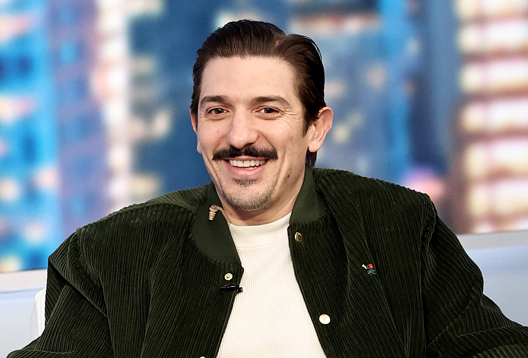 A idiosyncratic   smiling, seated indoors, wearing a corduroy overgarment   implicit    a airy  shirt. Blurred cityscape successful  the background