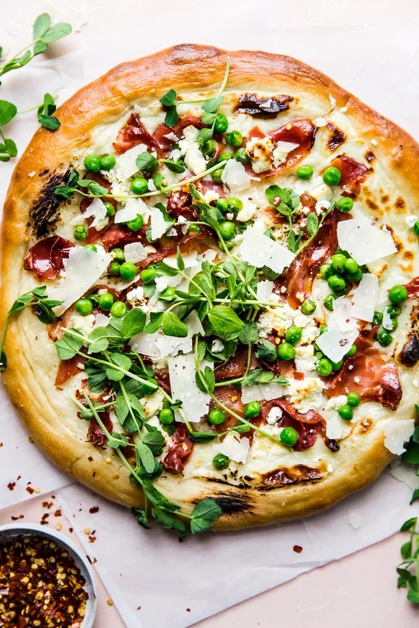 A gourmet pizza topped with prosciutto, greenish  peas, arugula, and parmesan shavings connected  a airy  crust